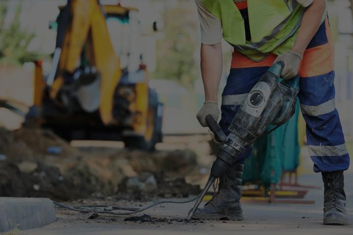 Pracownik w odblaskowej kamizelce używa młota pneumatycznego do rozbijania betonowej nawierzchni, w tle widoczna koparka.