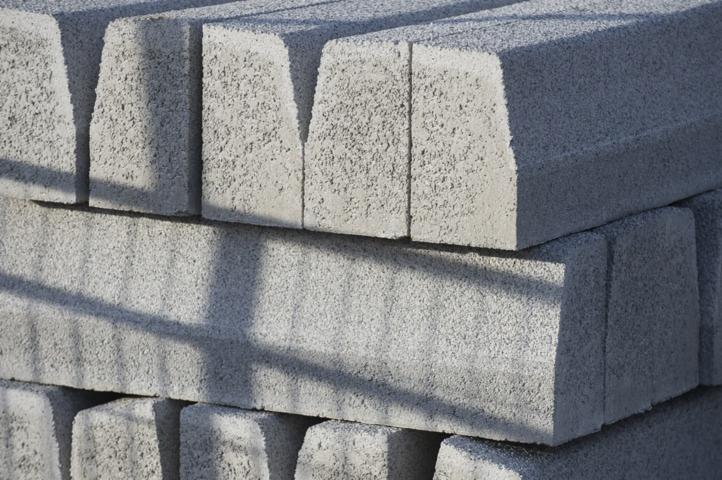 Stos szarych, betonowych obrzeży trawnikowych z charakterystycznym wycięciem, oświetlonych bocznym światłem słonecznym tworzącym wyraźny cień.