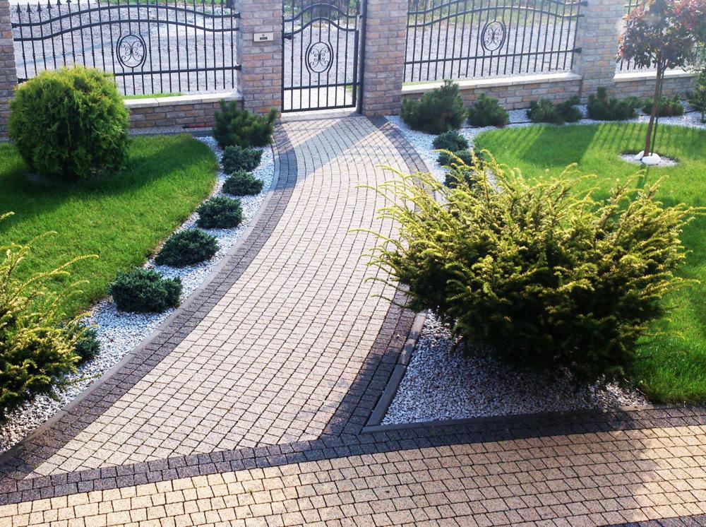 Ułożona z kostki brukowej ścieżka ogrodowa w kształcie łuku, obrzeża z ciemnej kostki, ozdobne krzewy i trawnik po bokach, brama z kutego żelaza w tle.