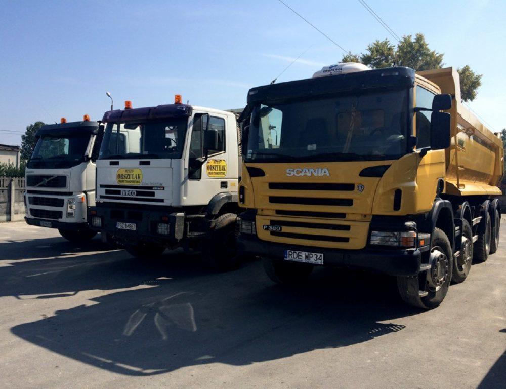 Trzy ciężarówki: żółta Scania P380 oraz białe Iveco i Volvo, zaparkowane na betonowym placu w słoneczny dzień.