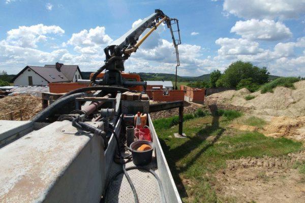 Betonomieszarka z wysięgnikiem w trakcie pracy na budowie domu jednorodzinnego, widok z góry na maszynę, w tle częściowo wzniesione mury z czerwonej cegły i hałda piasku.