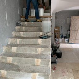 Styl Naprawa - Pracownik na schodach podczas przygotowywania podłoża do dalszych prac wykończeniowych, widoczne pojemniki z materiałami, narzędzia i surowe ściany.