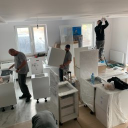 Styl Naprawa - Montaż białych szafek kuchennych: elektryk podłącza oświetlenie, stolarze składają korpusy, widoczne okna i elementy w folii ochronnej.