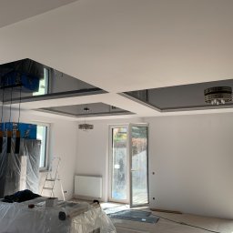 Styl Naprawa - Pokój w trakcie remontu z podwieszanym sufitem z elementami w kolorze grafitowym, lampami i oknami. Meble i sprzęty zabezpieczone folią, widoczne kartony i drabina.