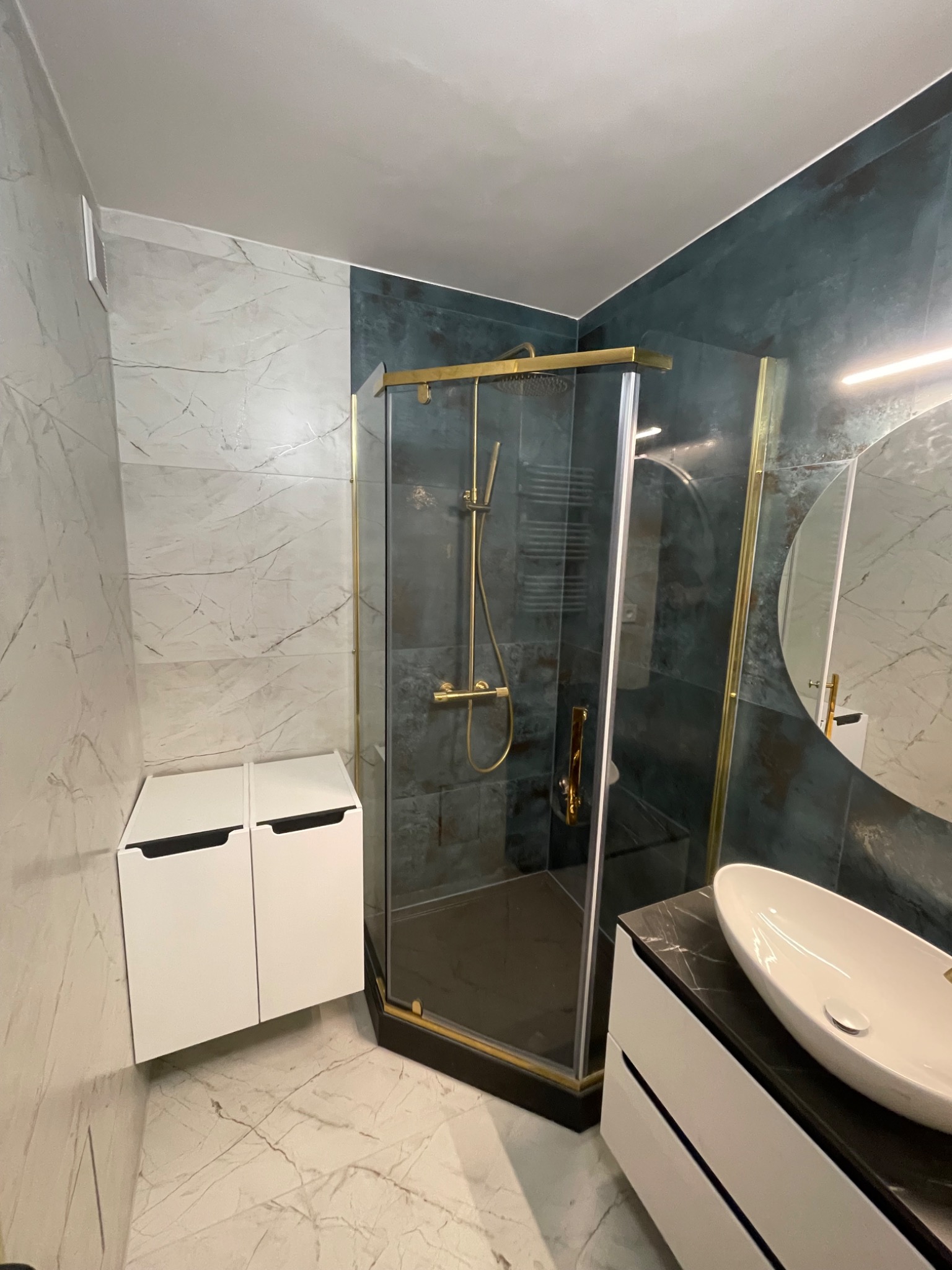 Nowoczesna łazienka z kabiną prysznicową w złotej ramie, umywalką nablatową na czarnym blacie i białymi szafkami, wykończona płytkami imitującymi marmur i ciemnymi płytkami na ścianie prysznica.