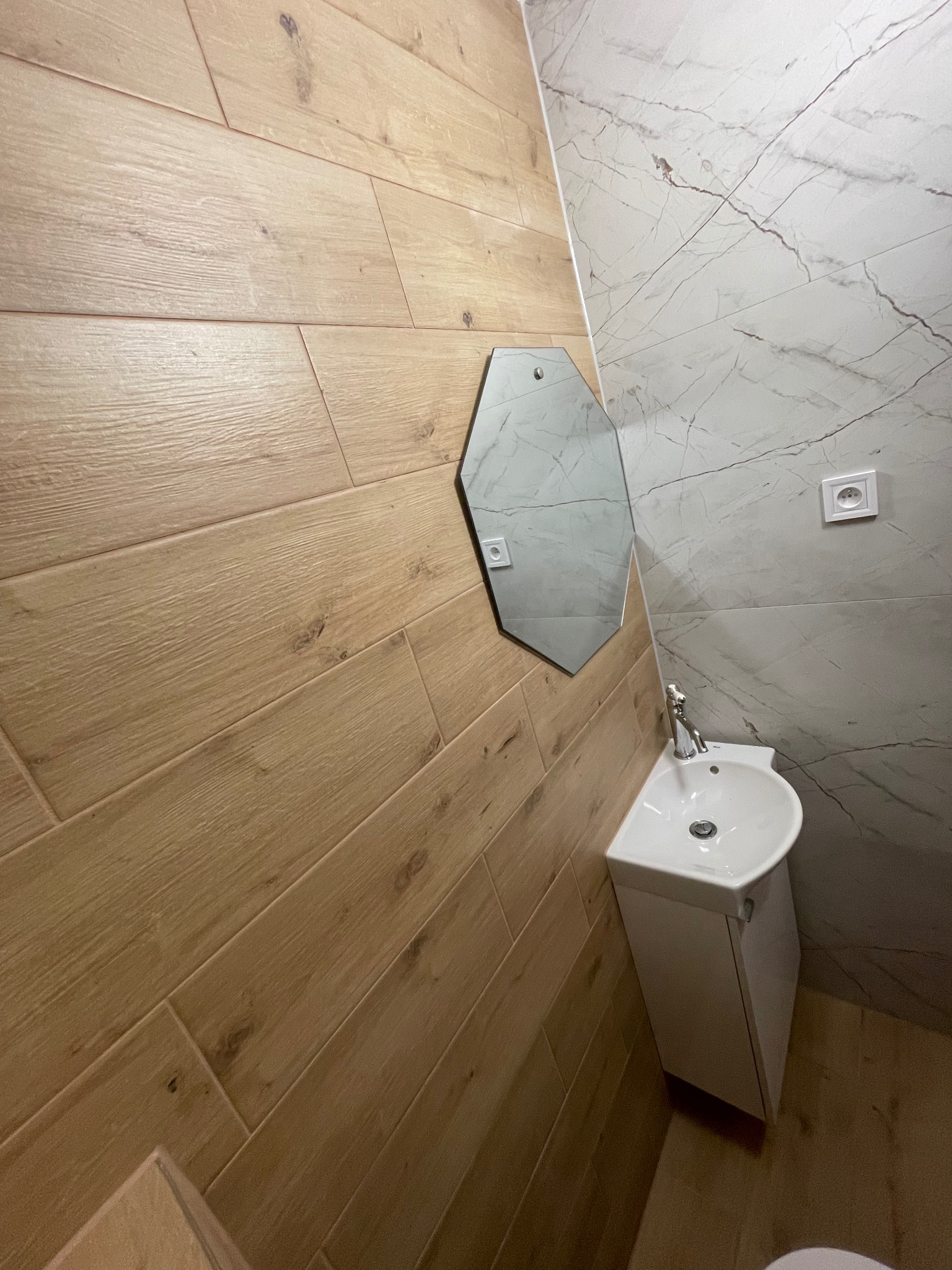 Mała łazienka z umywalką nablatową, heksagonalnym lustrem i okładzinami ściennymi imitującymi drewno oraz marmur.