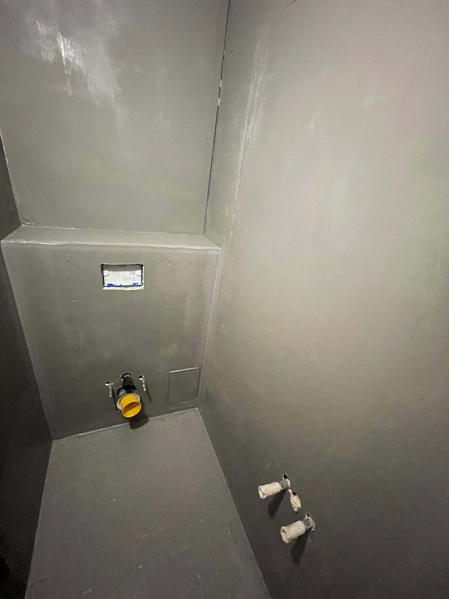 Przygotowana do położenia płytek, zagruntowana na szaro ściana w łazience z wyprowadzonymi instalacjami wodno-kanalizacyjnymi i otworem na puszkę elektryczną.