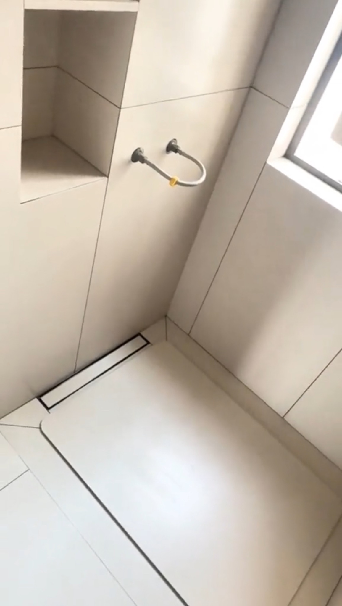 Nowoczesna łazienka z jasnymi, dużymi płytkami, widoczny odpływ liniowy, wnęka w ścianie i fragment rury doprowadzającej wodę z żółtą opaską.