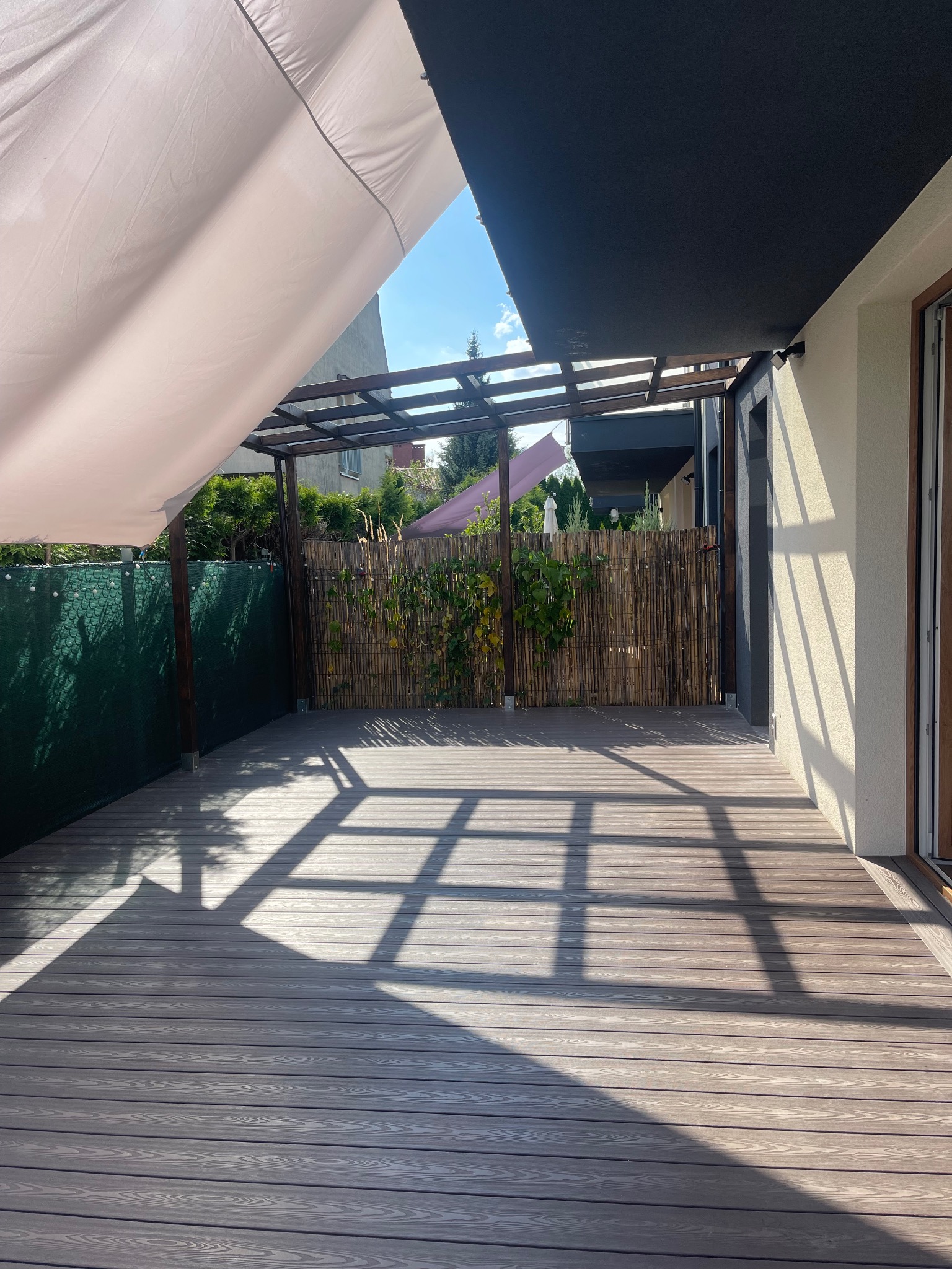 Drewniany taras z zadaszeniem pergolą, widoczne cienie konstrukcji, bambusowe ogrodzenie i fragment zielonego żywopłotu w tle.