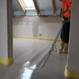 Pracownik w pomarańczowej kurtce rozprowadza płynną masę na podłodze w pomieszczeniu z oknem dachowym, widoczne żółte taśmy przy ścianach.