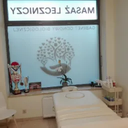 Wnętrze gabinetu z białym łóżkiem do zabiegów, na parapecie puchary, obok wózek z kosmetykami, okno z logo gabinetu.