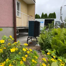 Zewnętrzna jednostka urządzenia grzewczego na metalowym stelażu, umieszczona przed budynkiem, z widocznym logotypem firmy instalacyjnej na samochodzie w tle.