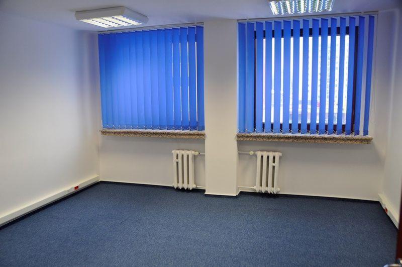 Puste biuro z niebieską wykładziną, dwoma oknami z pionowymi żaluzjami i białymi grzejnikami.