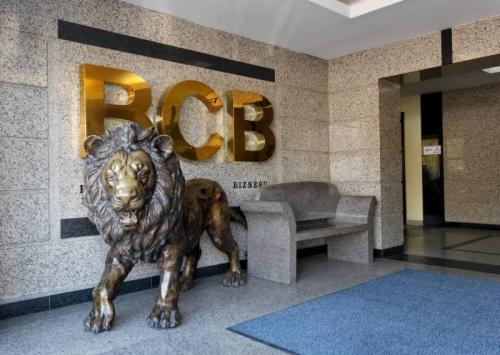 Lobby biurowca z marmurowymi ścianami, złotym napisem RCB BIZNESU, rzeźbą lwa i kamienną ławką.