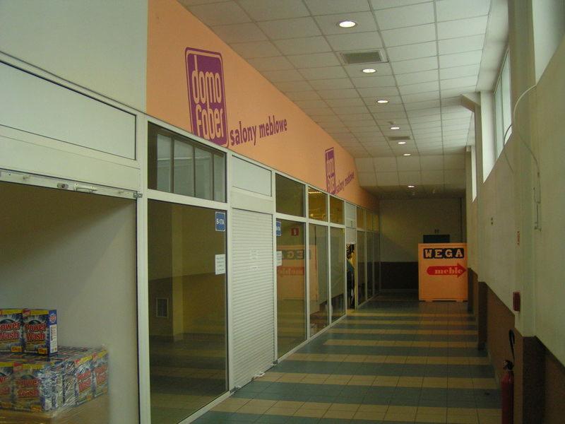 Wnętrze pasażu handlowego z lokalami do wynajęcia, widoczne witryny sklepowe z roletami, logo 'Domo Faber salony meblowe' na ścianie, paleta z towarem przed jednym z lokali.