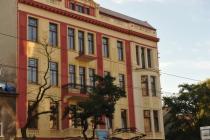 Elewacja odnowionego, trzypiętrowego budynku biurowego w stylu kamienicy, z balkonami z kutymi balustradami i czerwono-żółtą fasadą, widoczna z poziomu ulicy, z drzewami na pierwszym planie.