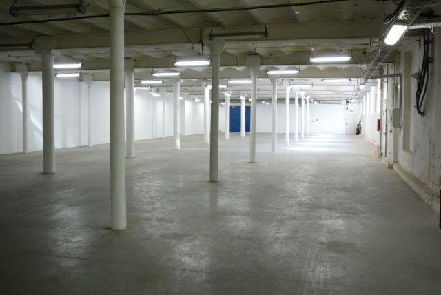 Wnętrze hali produkcyjnej lub magazynowej z betonową podłogą, rzędem białych kolumn, oświetleniem jarzeniowym pod sufitem i niebieskimi drzwiami w tle.