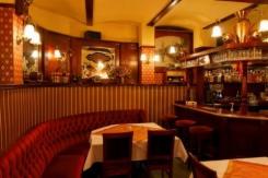 Wnętrze lokalu gastronomicznego w stylu retro: czerwona, tapicerowana kanapa w kształcie półkola, stolik z białym obrusem, bar z wysokimi stołkami. Na ścianach boazeria i dekoracyjne lustra. Uwagę...