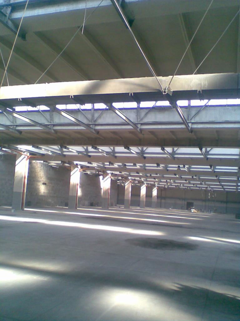 Wnętrze pustego magazynu z betonową podłogą, widoczne rzędy kolumn i konstrukcja dachu z oknami przepuszczającymi światło słoneczne, tworzące długie cienie na posadzce.