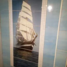 Błękitne płytki ścienne z motywem żaglowca, wkomponowane w ramę z małych, białych płytek. Widoczne niedokładności w ułożeniu fug.