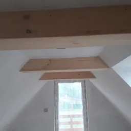 Poddasze z oknem dachowym i drewnianymi belkami stropowymi, ściany pomalowane na biało, widoczne ślady po szpachlowaniu i przygotowaniu do dalszych prac wykończeniowych.