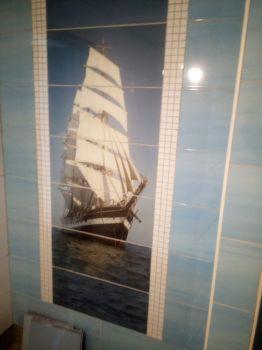 Błękitne płytki ścienne z motywem żaglowca, wkomponowane w ramę z małych, białych płytek. Widoczne niedokładności w ułożeniu fug.