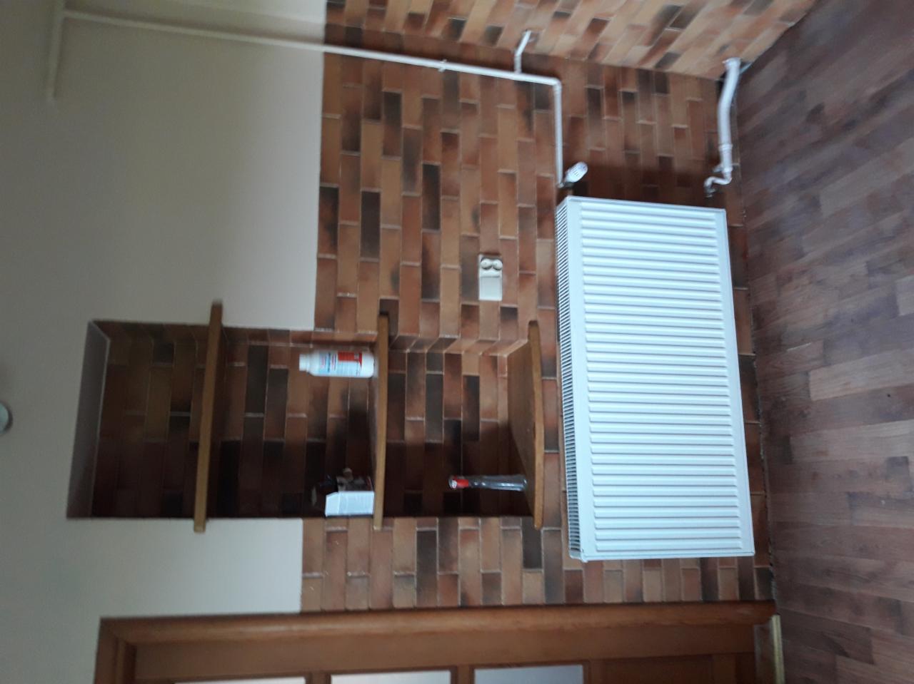 Wykończona ściana z brązowych płytek imitujących cegłę, biały grzejnik na ścianie, drewniana podłoga, wnęka w ścianie z drewnianymi półkami.