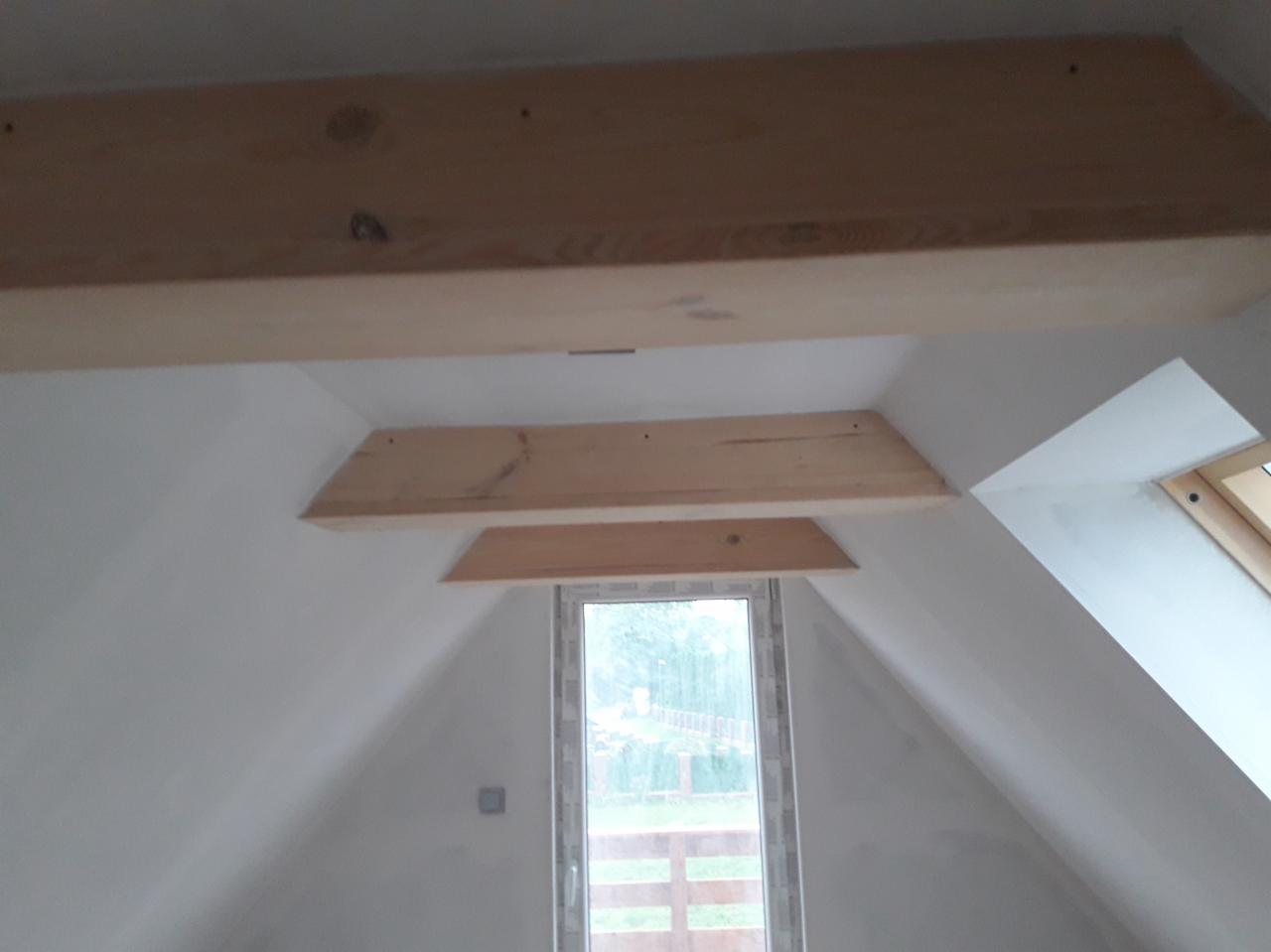 Poddasze z oknem dachowym i drewnianymi belkami stropowymi, ściany pomalowane na biało, widoczne ślady po szpachlowaniu i przygotowaniu do dalszych prac wykończeniowych.