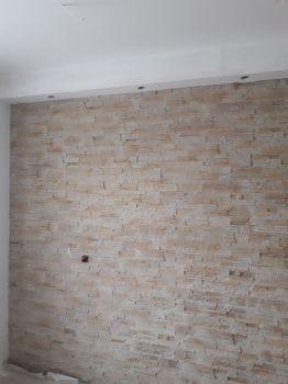 Ściana wyłożona dekoracyjnym kamieniem w odcieniach beżu i brązu, z widocznym gniazdkiem elektrycznym i surowym wykończeniem pod sufitem. Kamień ułożony w regularny wzór, tworząc teksturowaną...