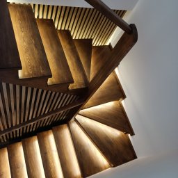 ZDOBRYMSMAKIEM.PL KRZYSZTOF RUTKOWSKI - Drewniane schody spiralne z podświetleniem LED, widok z dołu. Ciepłe światło podkreśla strukturę drewna i nowoczesny design. Minimalistyczne białe ściany w tle.