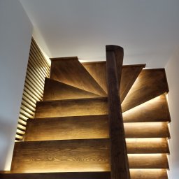ZDOBRYMSMAKIEM.PL KRZYSZTOF RUTKOWSKI - Drewniane schody spiralne z podświetleniem LED, widok z dołu, ukazujące detale słojów drewna i minimalistyczny design. Obok widoczna drewniana ściana z listew.
