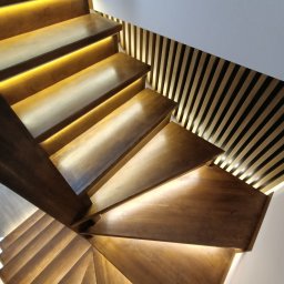 ZDOBRYMSMAKIEM.PL KRZYSZTOF RUTKOWSKI - Drewniane schody z podświetleniem LED i dekoracyjną ścianą z drewnianych listew, widok z góry, ciepłe światło.