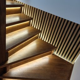 ZDOBRYMSMAKIEM.PL KRZYSZTOF RUTKOWSKI - Drewniane schody z podświetleniem LED, widok z dołu. Obok ażurowa ścianka z drewnianych listewek. Ciemne drewno stopni kontrastuje z jasnym tłem.