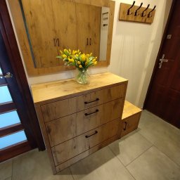 ZDOBRYMSMAKIEM.PL KRZYSZTOF RUTKOWSKI - Drewniana szafka z lustrem i wazonem żółtych tulipanów w przedpokoju. Nad nią wieszak na ubrania. Całość w ciepłej tonacji, z widoczną strukturą drewna.