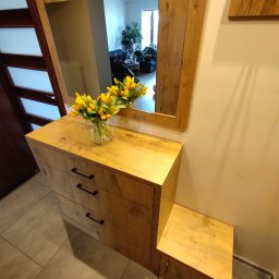 ZDOBRYMSMAKIEM.PL KRZYSZTOF RUTKOWSKI - Drewniana komoda z trzema szufladami i lustrem w ramie z tego samego drewna. Na komodzie wazon z żółtymi tulipanami. Widoczny wieszak na ścianie i fragment drzwi.