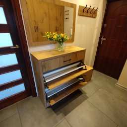 ZDOBRYMSMAKIEM.PL KRZYSZTOF RUTKOWSKI - Drewniana szafka na buty z lustrem i wazonem tulipanów w przedpokoju. Widoczne drzwi wejściowe i wieszak na ubrania. Ujęcie z dołu, podkreślające przestrzeń.