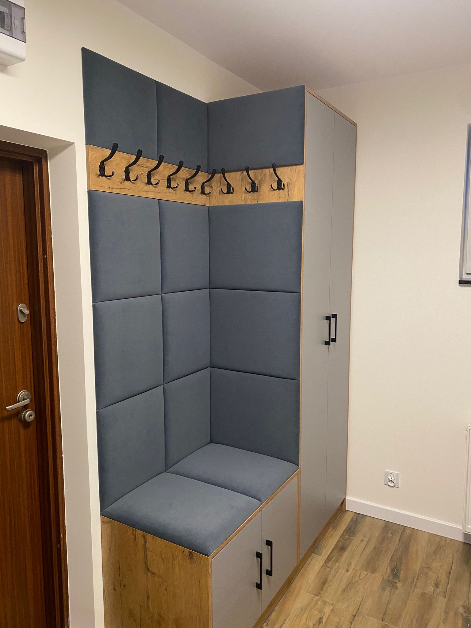 Szara zabudowa przedpokoju z miękkim siedziskiem i wieszakami, drewniane wykończenie, szara szafa obok. Nowoczesny design.