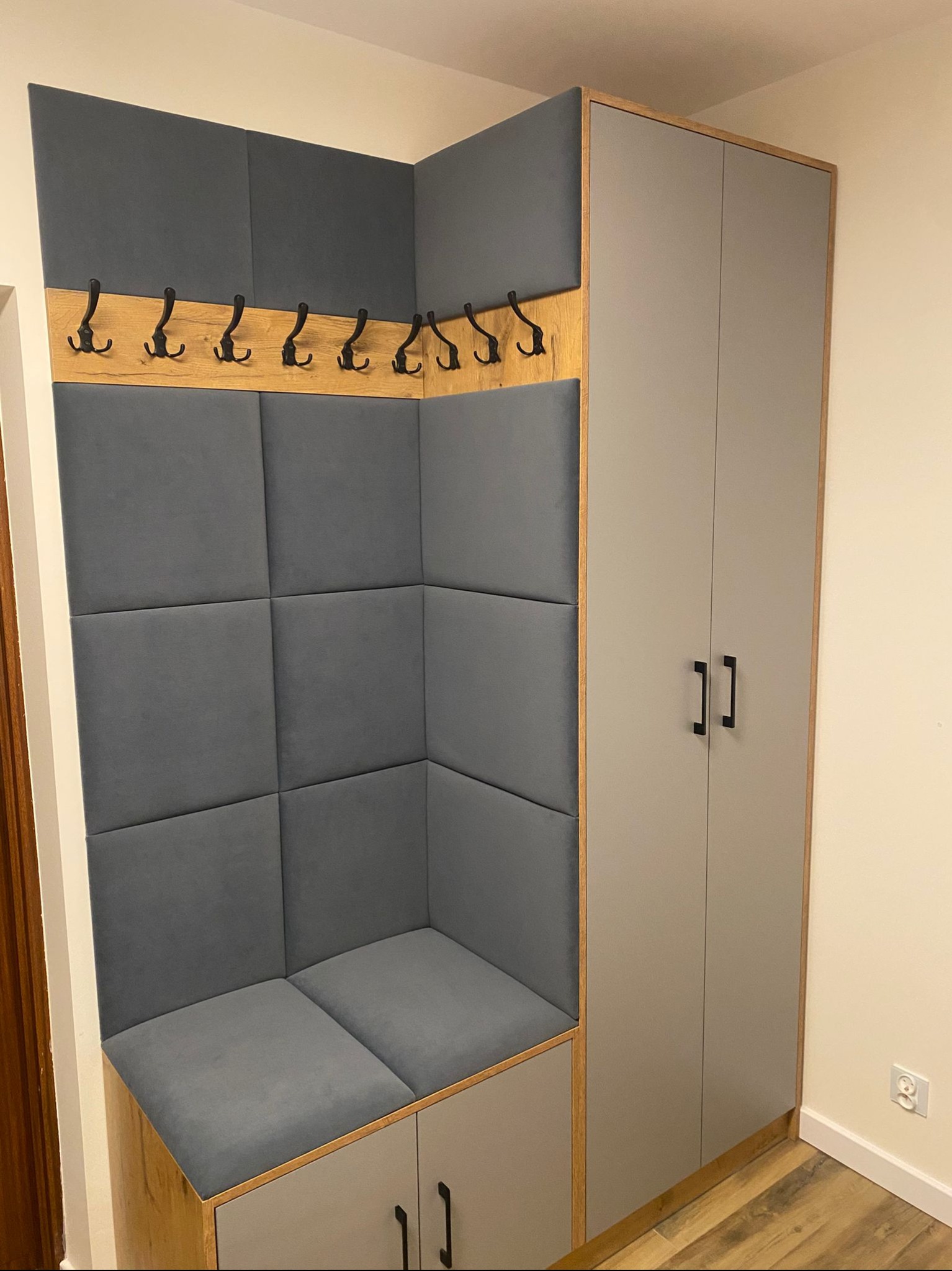 Szara zabudowa przedpokoju z siedziskiem i wieszakami, obok szafa z drewnianymi obramowaniami. Nowoczesny design, neutralna kolorystyka, praktyczne rozwiązanie do przechowywania.