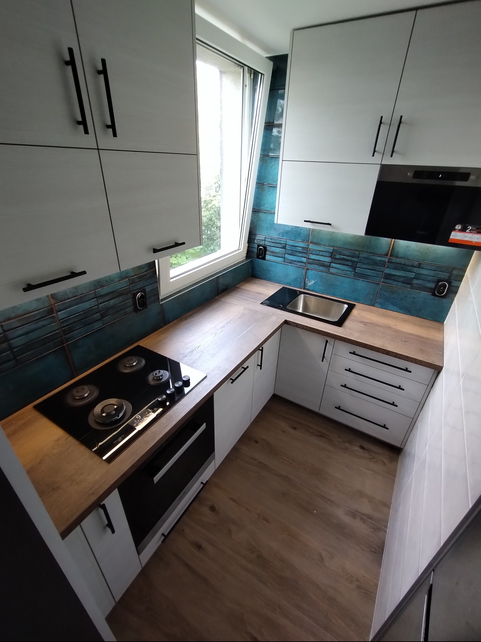 Nowoczesna kuchnia w bieli i drewnie z turkusowymi płytkami. Ujęcie z góry podkreśla ergonomiczny układ szafek i sprzętów, w tym płyty grzewczej i zlewu.