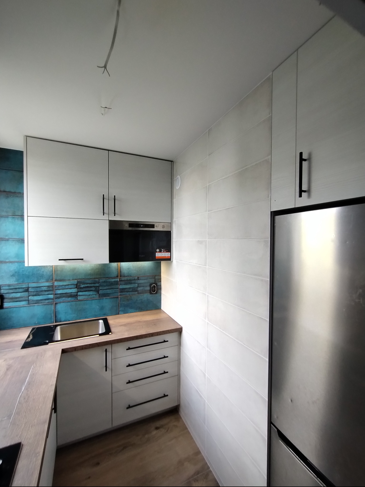 Nowoczesna kuchnia z białymi szafkami i turkusowymi płytkami. Stalowa lodówka i drewniany blat dodają elegancji. Widoczne niedokończone kable pod sufitem.