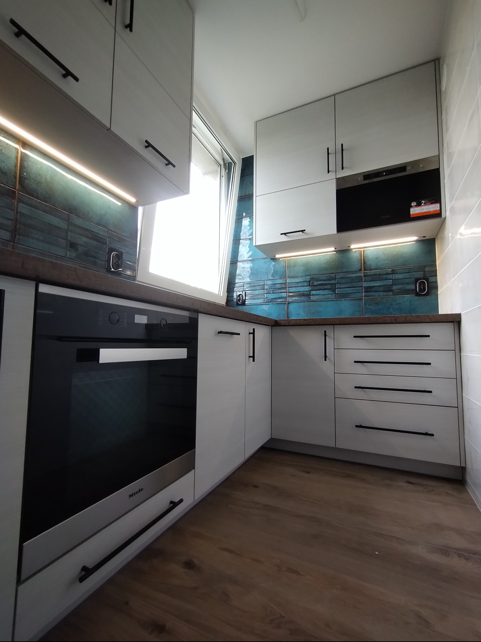 Biała kuchnia z czarnymi uchwytami, oświetleniem LED podszafkowym i turkusowymi płytkami nad blatem. Widoczny piekarnik marki Miele i drewniana podłoga.