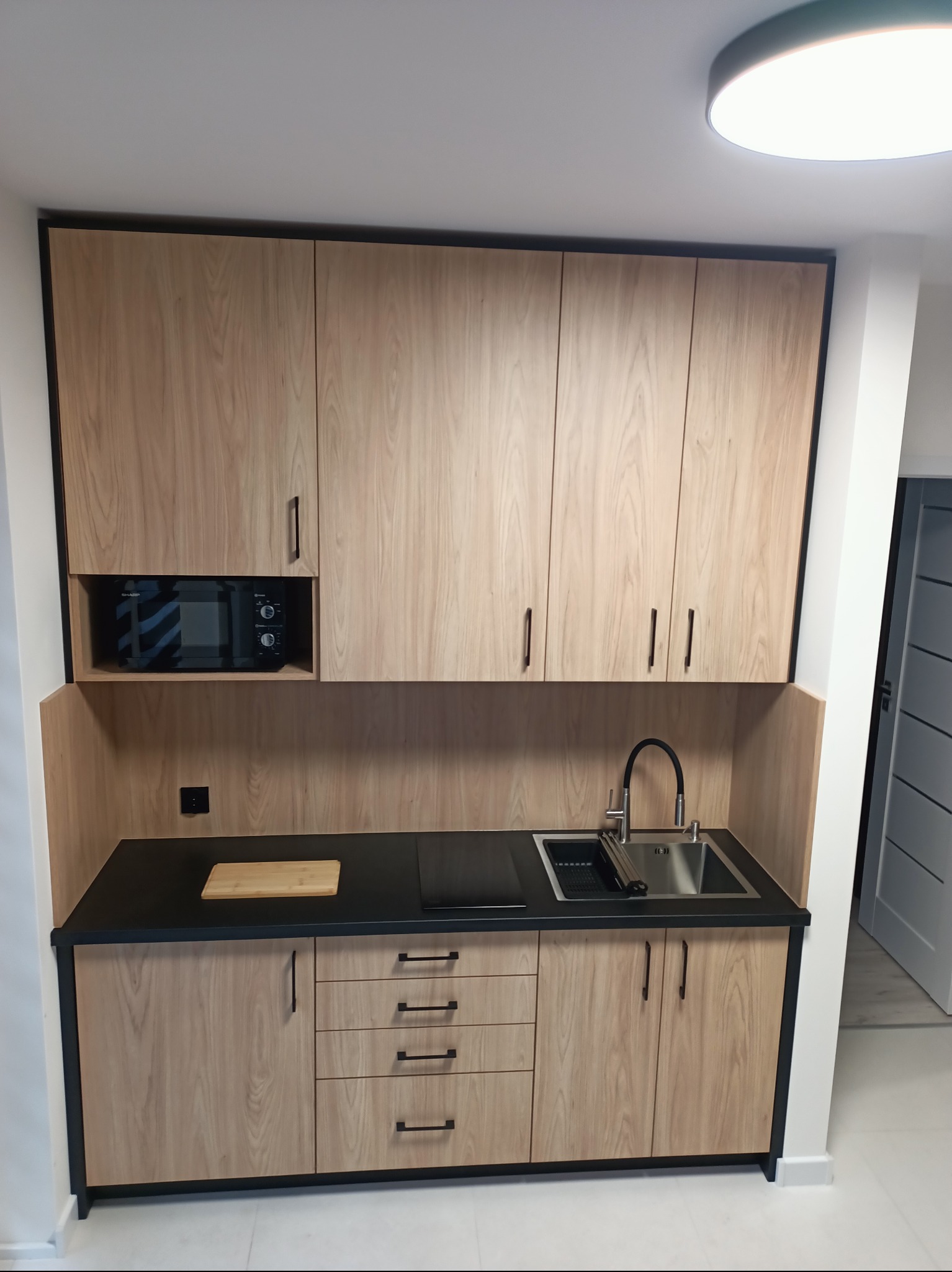 Jasna kuchnia na wymiar z czarnym blatem i ramami szafek, z wbudowaną mikrofalówką i deską do krojenia, minimalistyczny design.