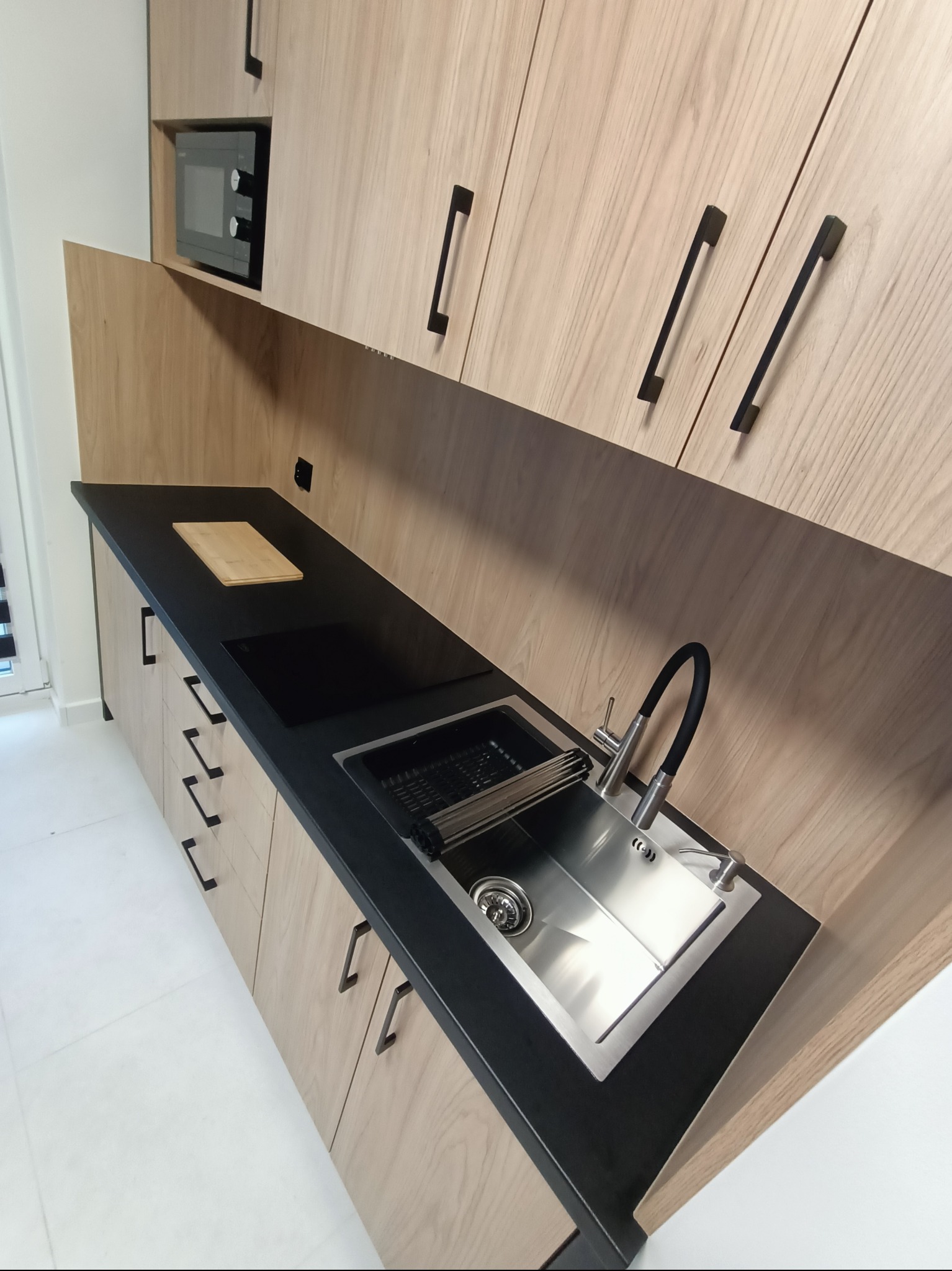 Jasna kuchnia na wymiar z drewnianymi frontami, czarnym blatem i metalowym zlewem. Widoczna płyta indukcyjna i deska do krojenia.