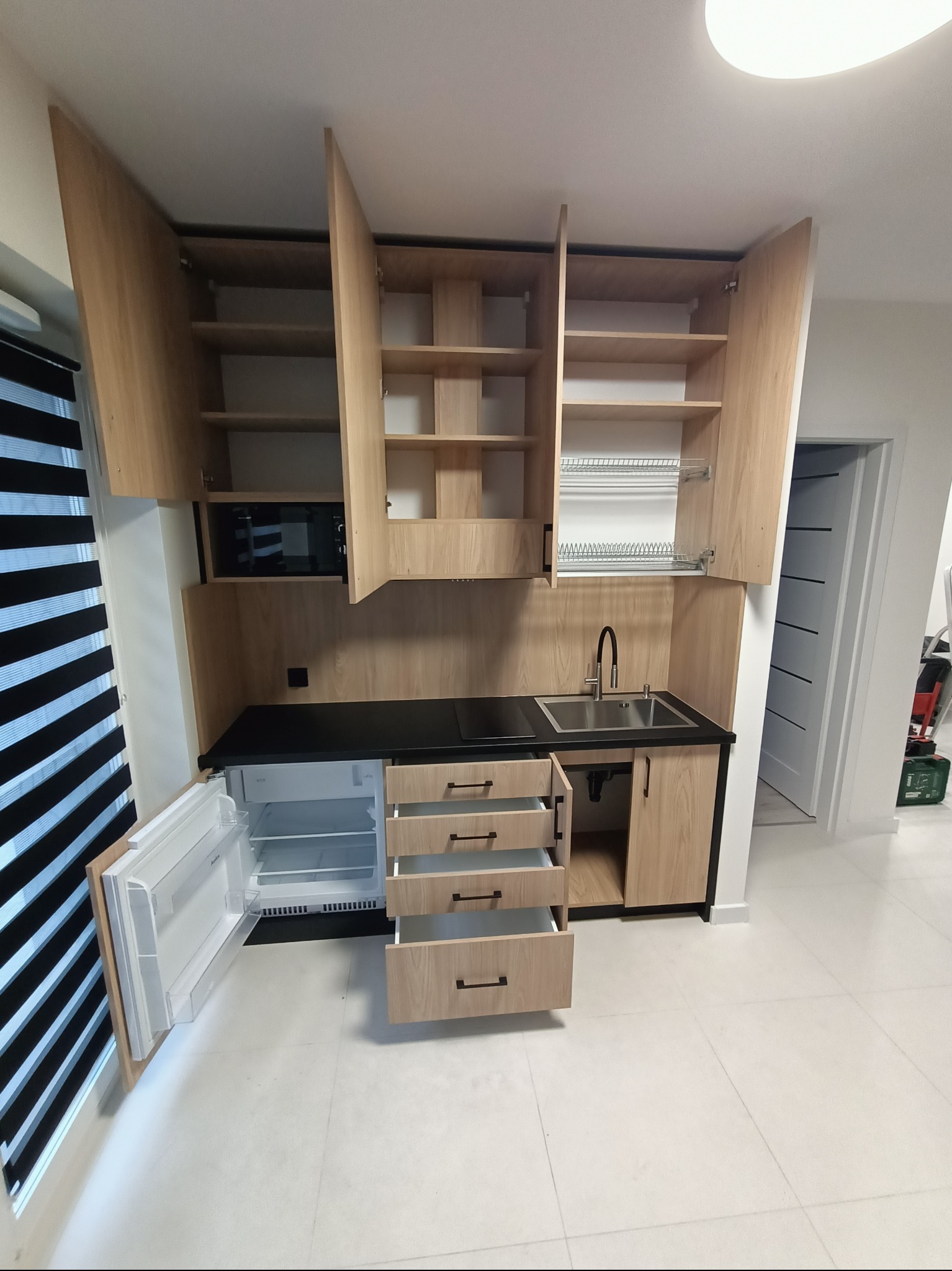 Jasna zabudowa kuchenna z drewnianymi frontami, czarnym blatem i otwartymi szafkami, mała lodówka pod blatem, nowoczesny design.