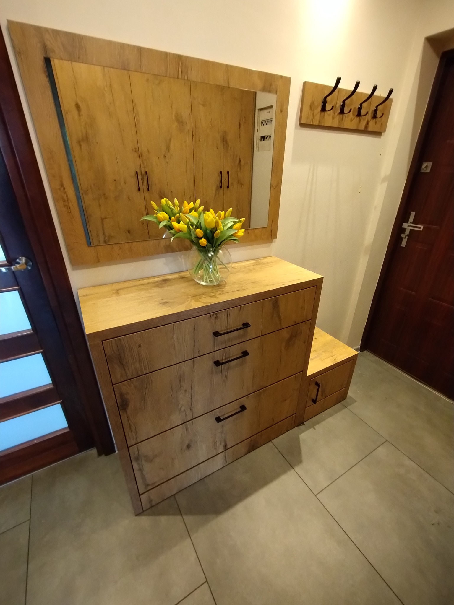 Drewniana szafka z lustrem i wazonem żółtych tulipanów w przedpokoju. Nad nią wieszak na ubrania. Całość w ciepłej tonacji, z widoczną strukturą drewna.