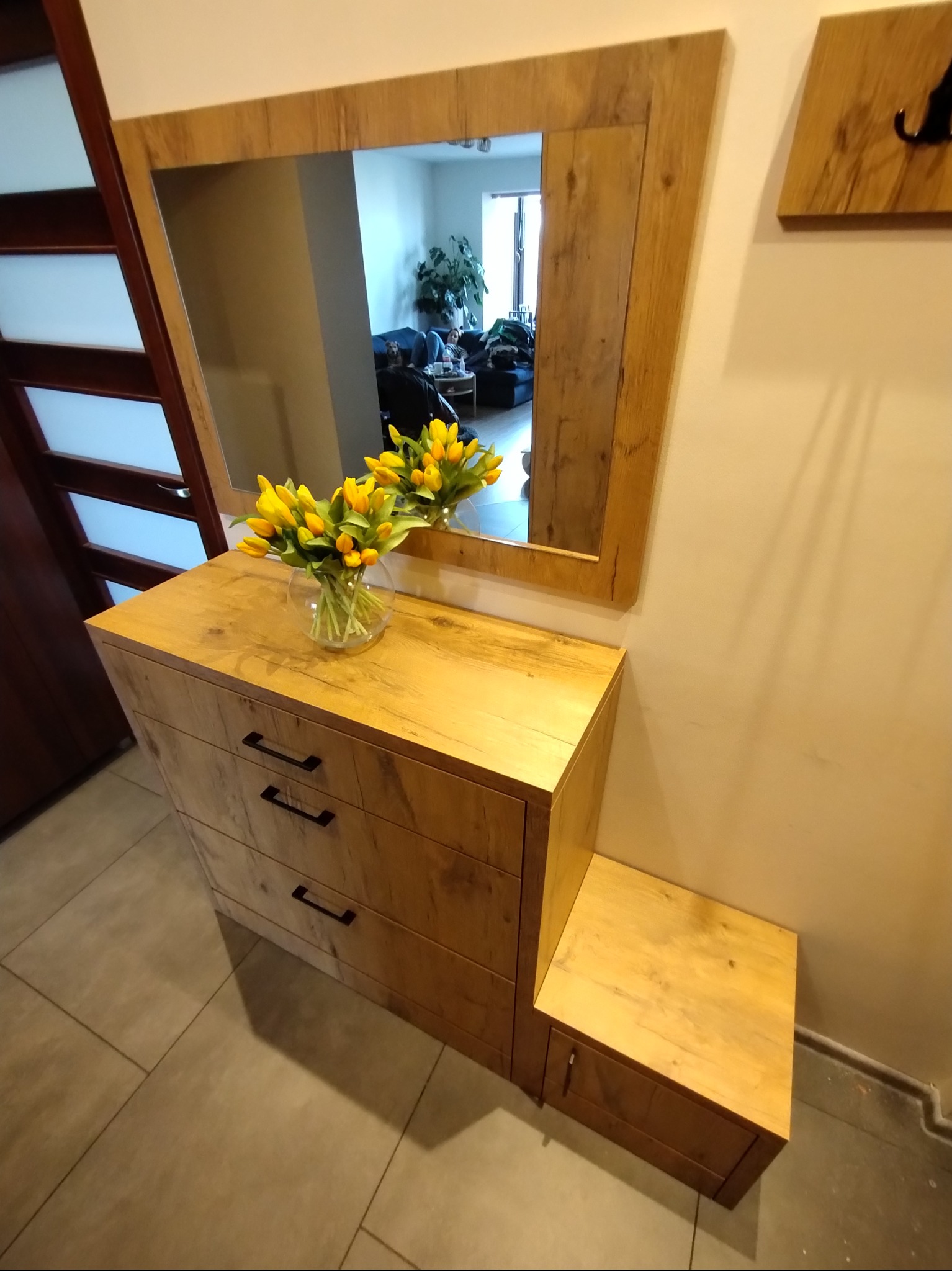 Drewniana komoda z trzema szufladami i lustrem w ramie z tego samego drewna. Na komodzie wazon z żółtymi tulipanami. Widoczny wieszak na ścianie i fragment drzwi.