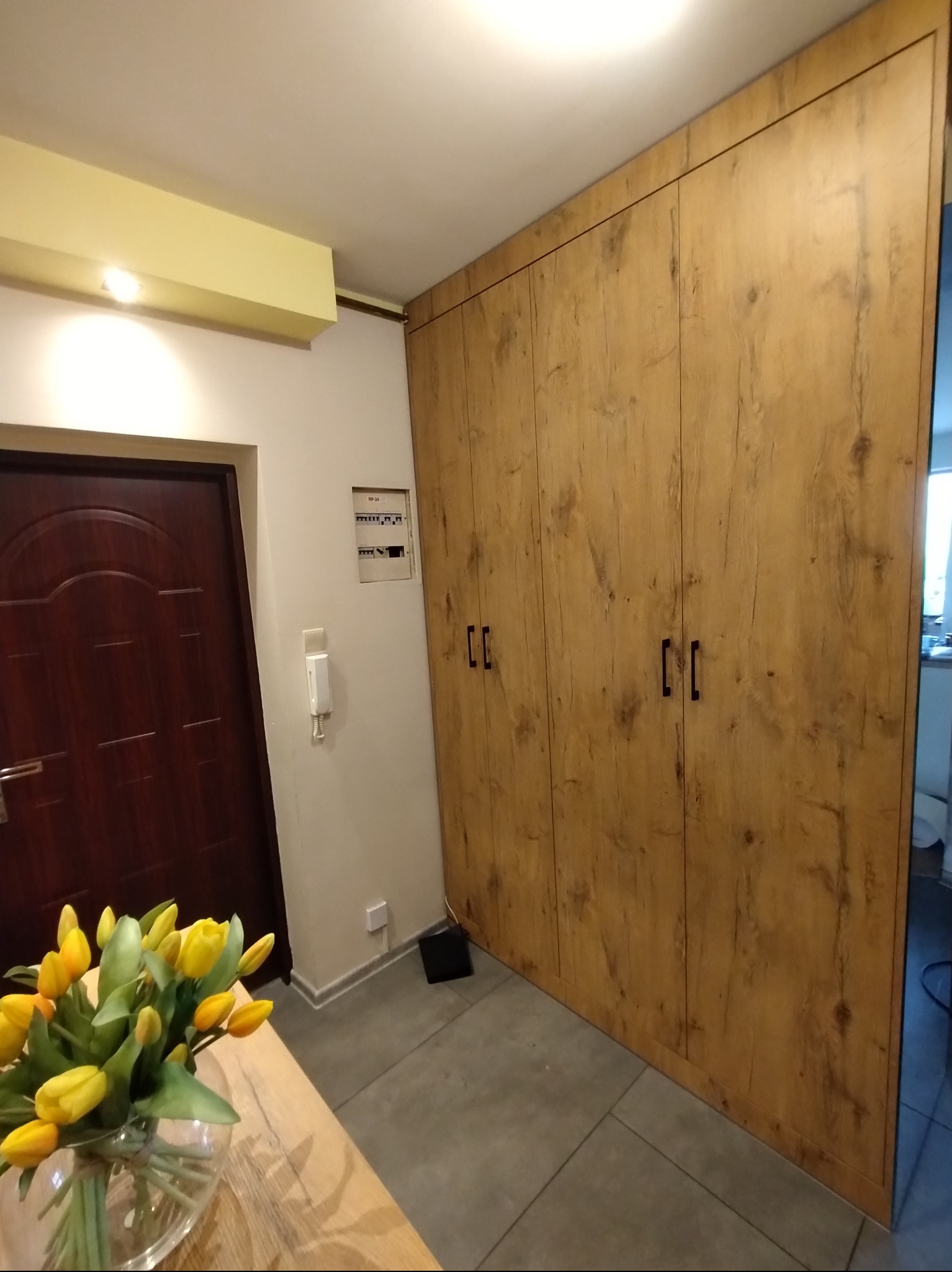 Drewniana szafa wnękowa z czterema drzwiami, czarne uchwyty, szara podłoga, widok z perspektywy, bukiet tulipanów w wazonie na pierwszym planie.