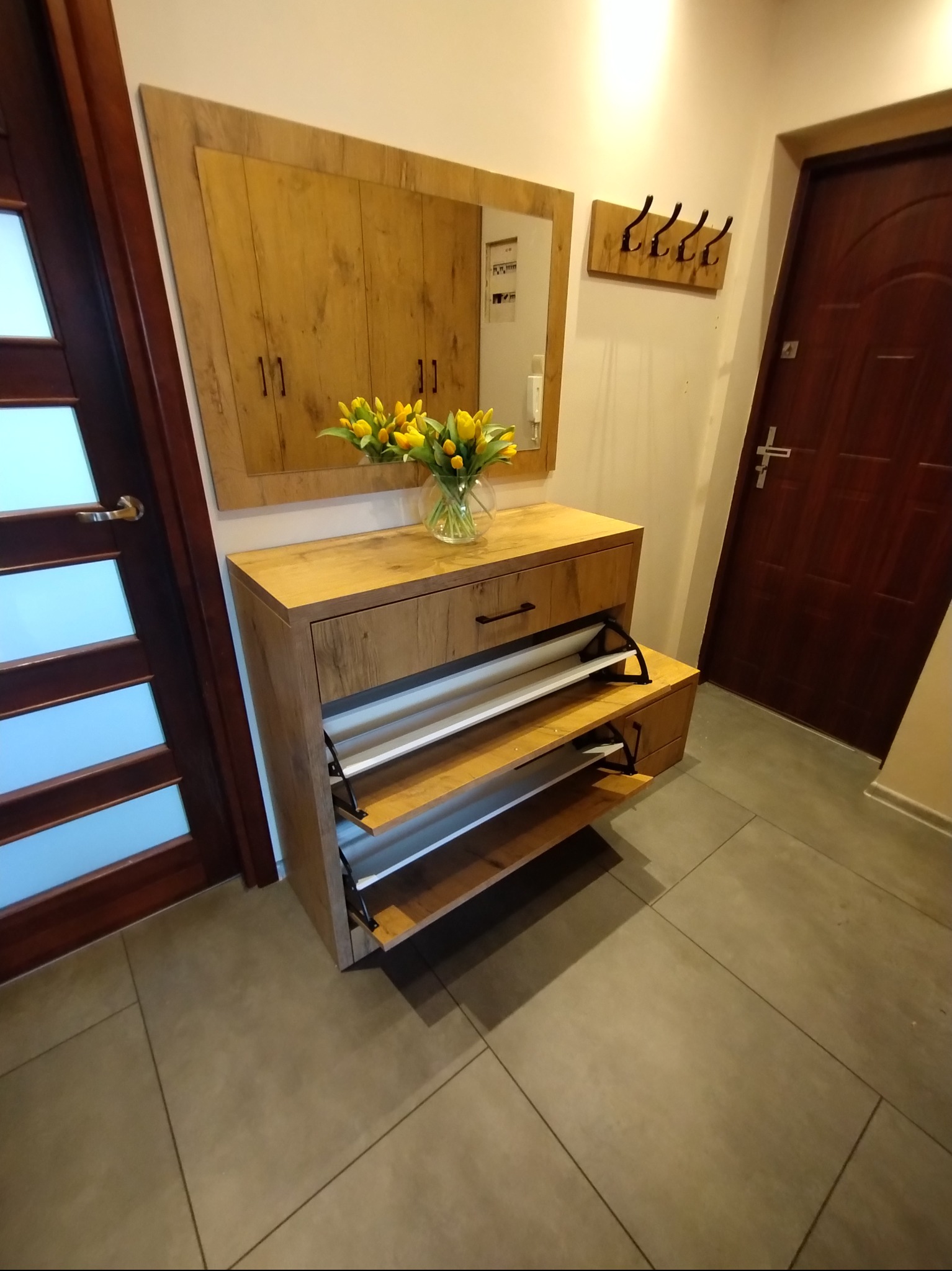 Drewniana szafka na buty z lustrem i wazonem tulipanów w przedpokoju. Widoczne drzwi wejściowe i wieszak na ubrania. Ujęcie z dołu, podkreślające przestrzeń.