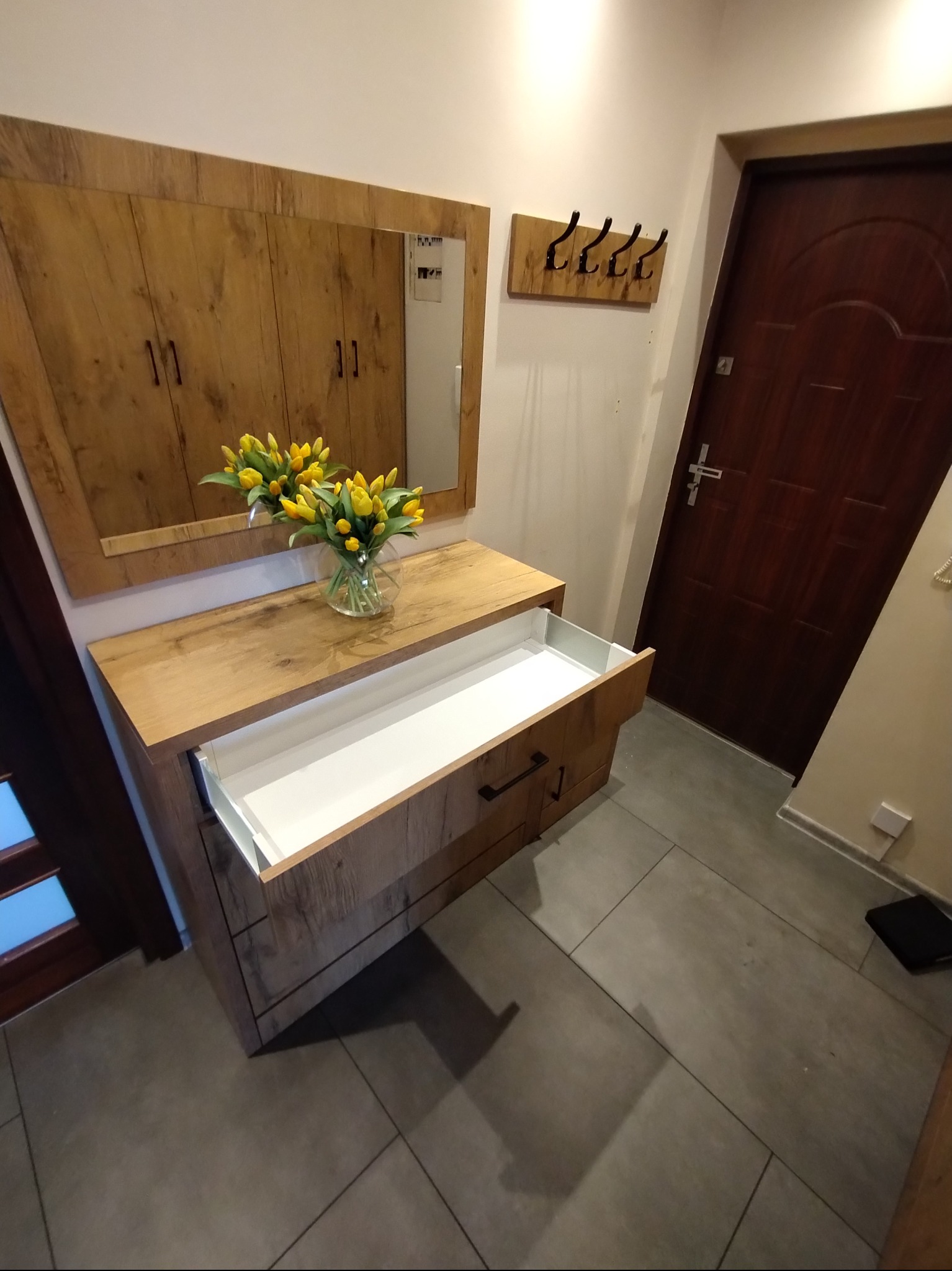 Drewniana szafka z lustrem i wysuwaną szufladą w przedpokoju, dekorowana żółtymi tulipanami w wazonie. Widoczny wieszak na ścianie i drzwi wejściowe.