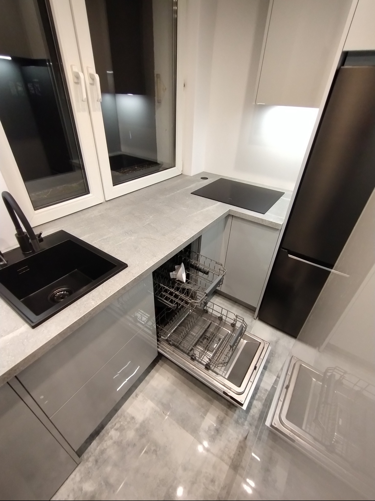 Szara, nowoczesna kuchnia z czarnym zlewem i otwartą zmywarką. Widoczne okno i płyta indukcyjna. Minimalistyczny design, błyszczące fronty szafek.